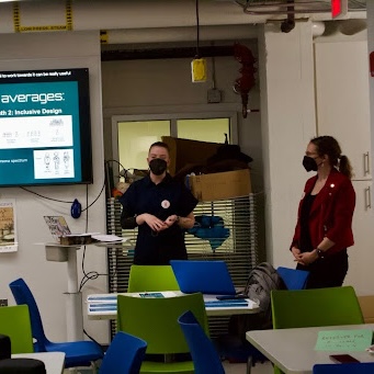 Three people collaborate at a table in a brightly lit, creative workspace. One person is bent over writing on a large sheet of paper, while two others stand nearby discussing and reviewing materials. The room features colorful chairs, bean bags, and murals on the walls, with a whiteboard and various supplies visible in the background.
