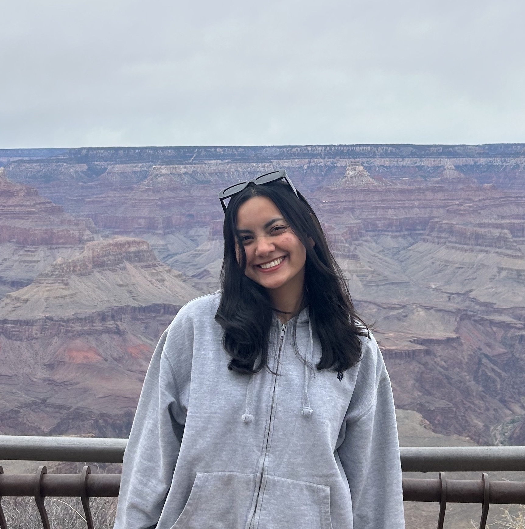 Babita, a brown girl with wavy black hair, smiles warmly at the camera with her head slightly tilted. She wears black sunglasses on top of her head and a grey zip-up jacket. The background features a black railing with the vast, scenic canyons of Arizona’s Grand Canyon.