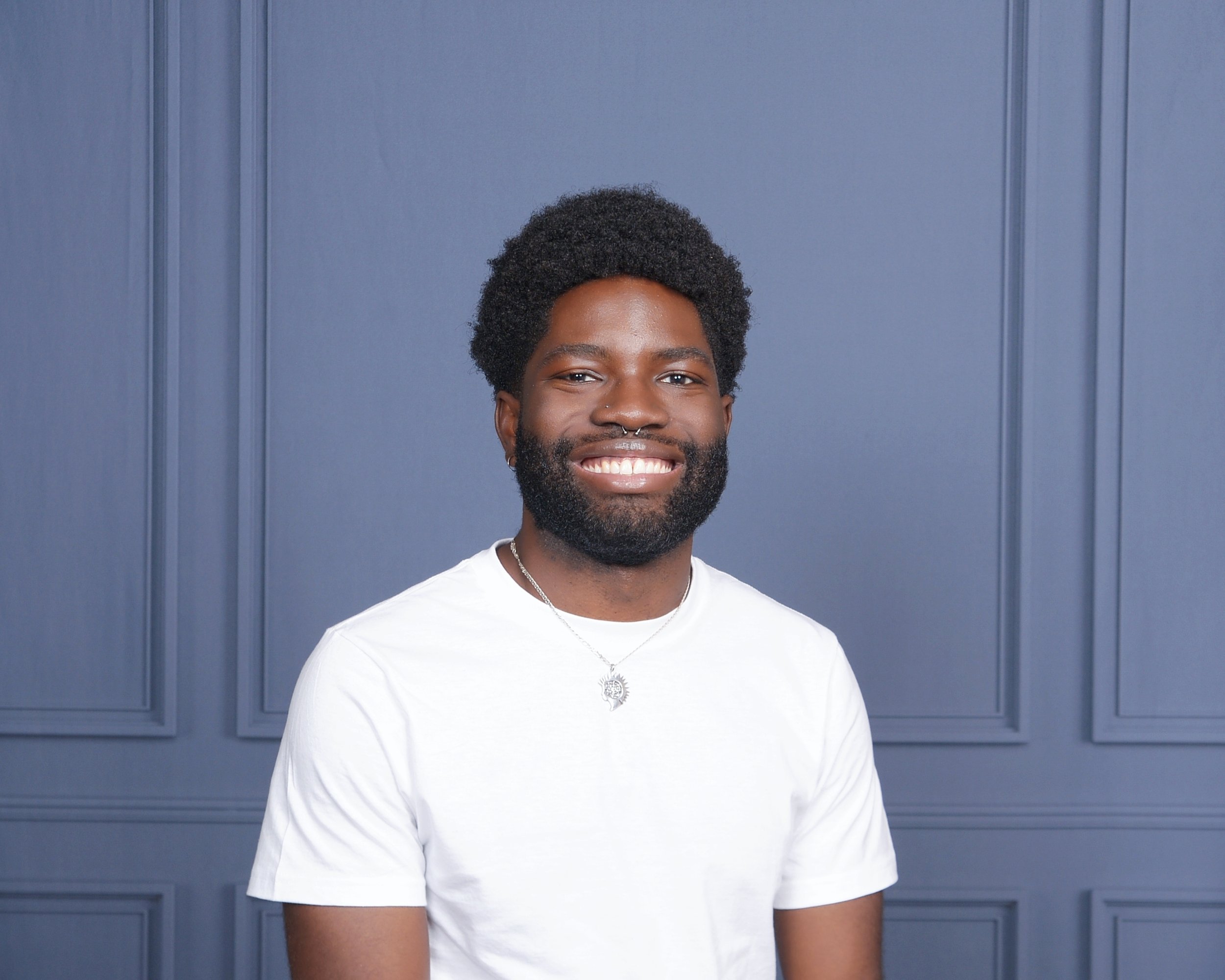 Bonsu, a Black man with a beard, a septum piercing, natural hair wearing a white t-shirt and necklace, standing in front of a blue-gray paneled wall facing to the right.
