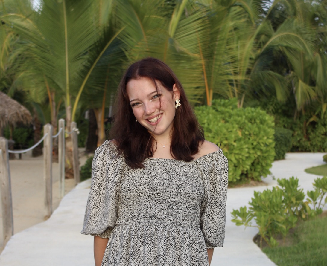 Julie, a white girl smiling widely with her head slightly tilted and her arms behind her back. She has shoulder length, wavy brown hair, which is pushed behind her ears with one piece falling in front of her face. She is wearing a small pendant necklace, polka-dotted earrings, and a spotted dress, standing in front of a vibrantly green background filled with palm trees.