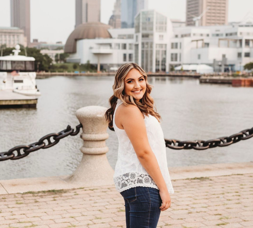 Karlynn, a white girl facing towards the right with her arms at her side and her head turned towards the camera, smiling wide. She has curled brown hair with blonde highlights, and is wearing a long, white lace tank top with dark blue jeans. The background shows a tan, brick sidewalk that gives way to blue water in a harbor. There is a large chain barrier that separates the land from the water. In the water, there are boats and docks. And beyond that are tall brown, blue, and white buildings of different shapes and sizes.