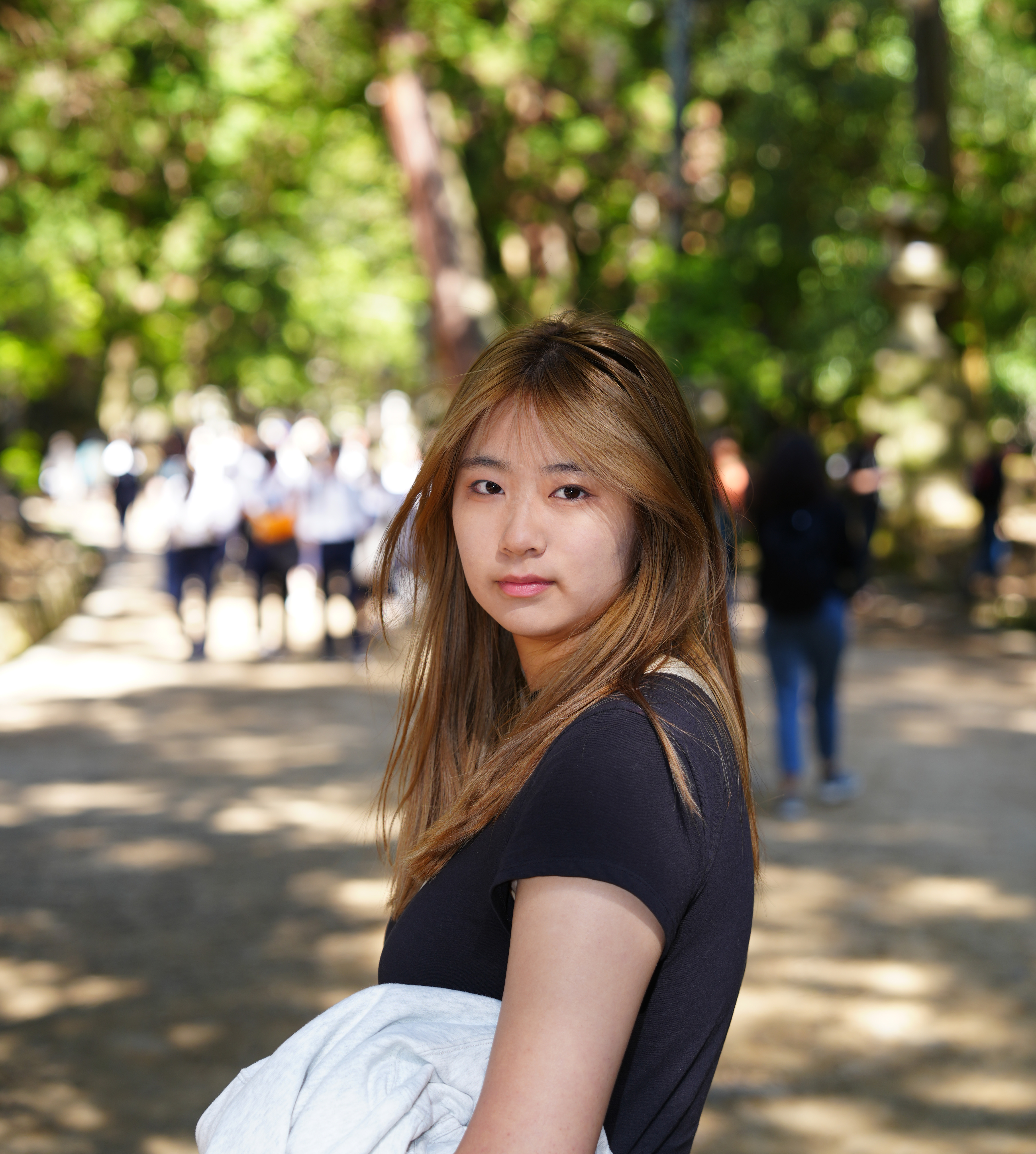 Kaylynn, an Asian girl standing on a sunlit forest path, facing to the right with her head turned toward the camera, wearing a calm and natural expression. She has straight, light brown hair and is dressed in a black short-sleeve top, holding a light-colored jacket in her arms. The background shows a tree-lined walkway dappled with sunlight, with a group of students in uniforms walking in the distance, creating a peaceful and bright outdoor atmosphere.