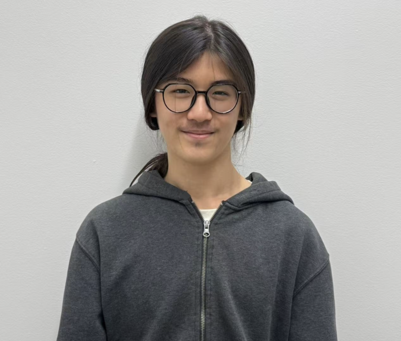 Peter, an Asian person with dark, long hair, wearing round black glasses and a charcoal‑gray zip‑up hoodie, giving a slight smile against a plain light‑gray background.