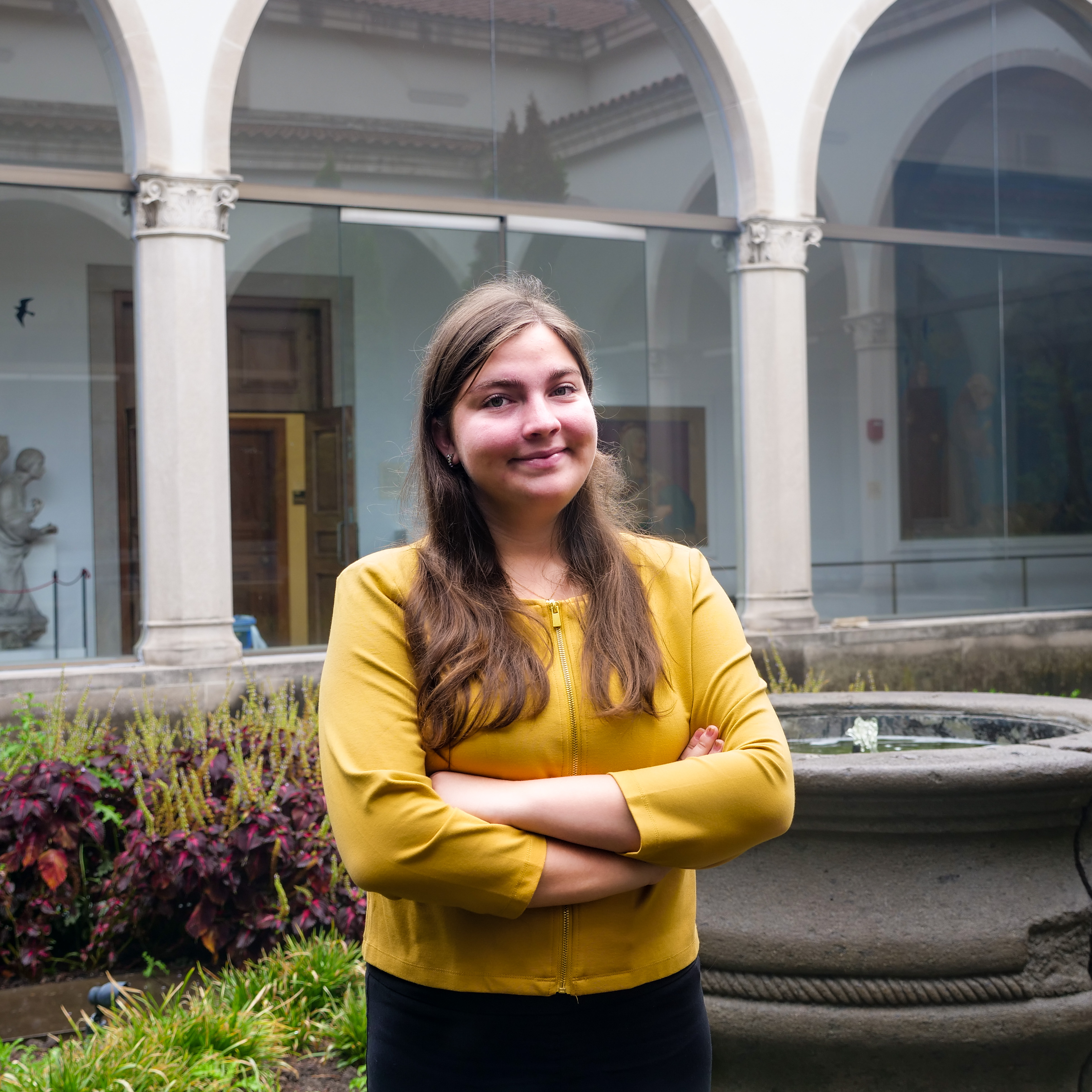 Tatyana, a white person with long, wavy brown hair. She is smiling softly with her arms crossed and looking at the camera, wearing a mustard-yellow zip-up jacket with black pants. The background showcases a courtyard with stone arches, glass panels reflecting greenery, and a fountain surrounded by plants, suggesting a museum or historical site.
