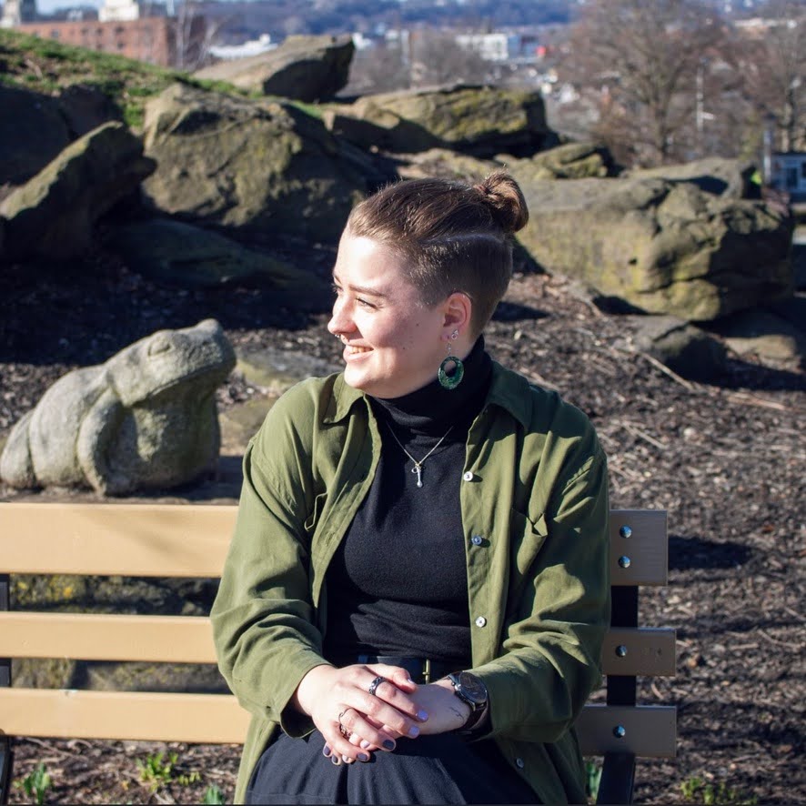 Lynn, a white person with their light brown hair in a top knot and a fade around the sides. They have a septum piercing and are smiling widely, looking off to the left. They are wearing a green button down shirt, a necklace with a spoon charm, and green computer chip earrings.