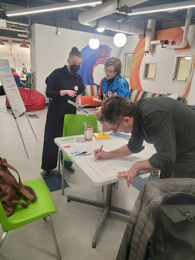 Three people collaborate at a table in a brightly lit, creative workspace. One person is bent over writing on a large sheet of paper, while two others stand nearby discussing and reviewing materials. The room features colorful chairs, bean bags, and murals on the walls, with a whiteboard and various supplies visible in the background.
