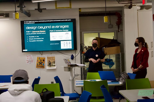 Lynn, a white person in all black with a black mask and Dr. FitzPatrick, a white woman wearing a red blazer with a black shirt and black mask giving a lecture at the front of a classroom.