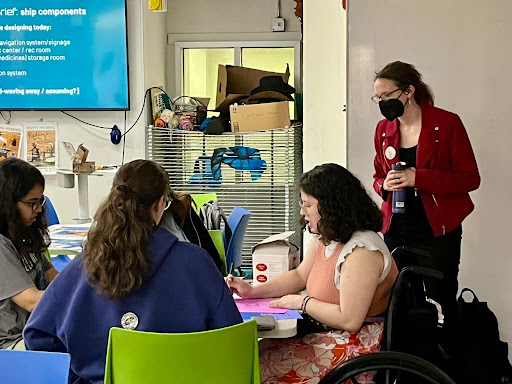 A group of people sit around a table working on the D.I.S.C.O. workshop, with one person using a wheelchair actively writing on pink paper. Another person in a red jacket and mask stands nearby, observing the working group. The room is filled with colorful chairs, a large screen displaying project instructions, and various supplies and equipment on shelves in the background.
