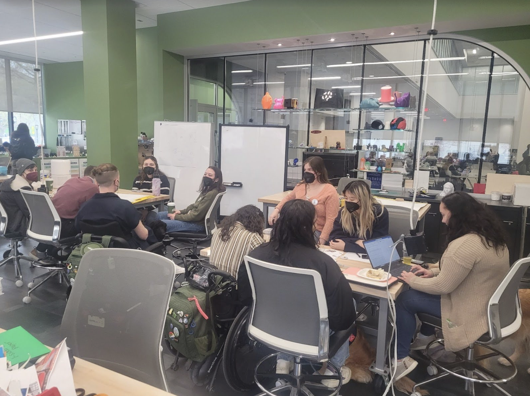 A group of people, two of them using wheelchairs, are gathered around tables in a collaborative workspace. They are wearing face masks and appear engaged in discussion or working on laptops. The room has green walls, whiteboards, and a glass wall showing a modern makerspace with 3D printers and various colorful models on display.